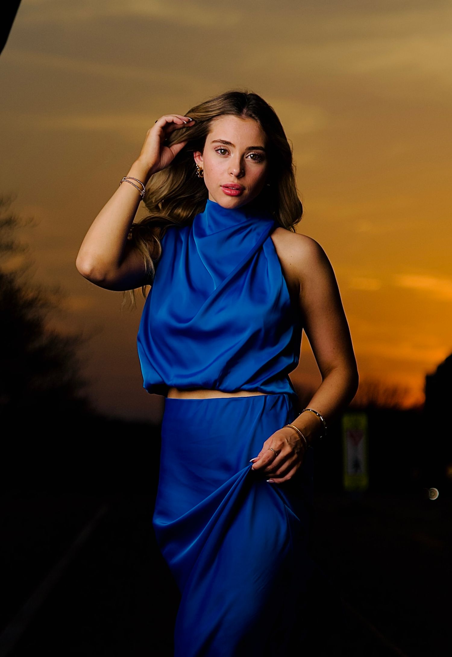 A figure in a vibrant blue dress poses dramatically against a golden sunset sky in a creative fashion photoshoot series.