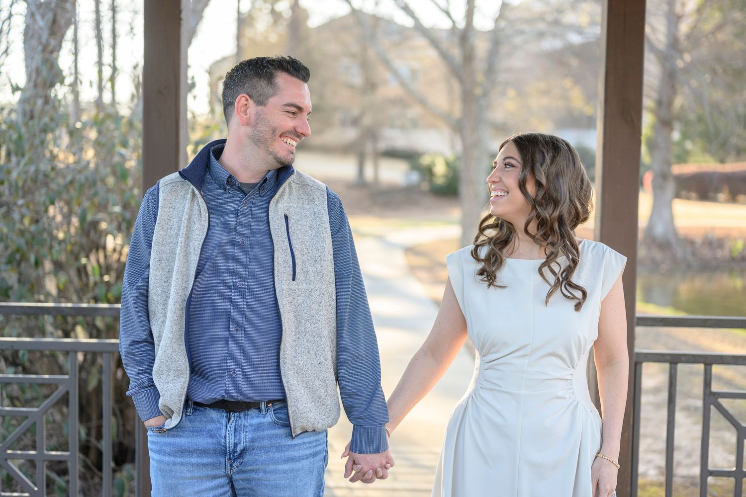 A couple wearing coordinated blue and white outfits smile at each other while standing outdoors in a park setting.