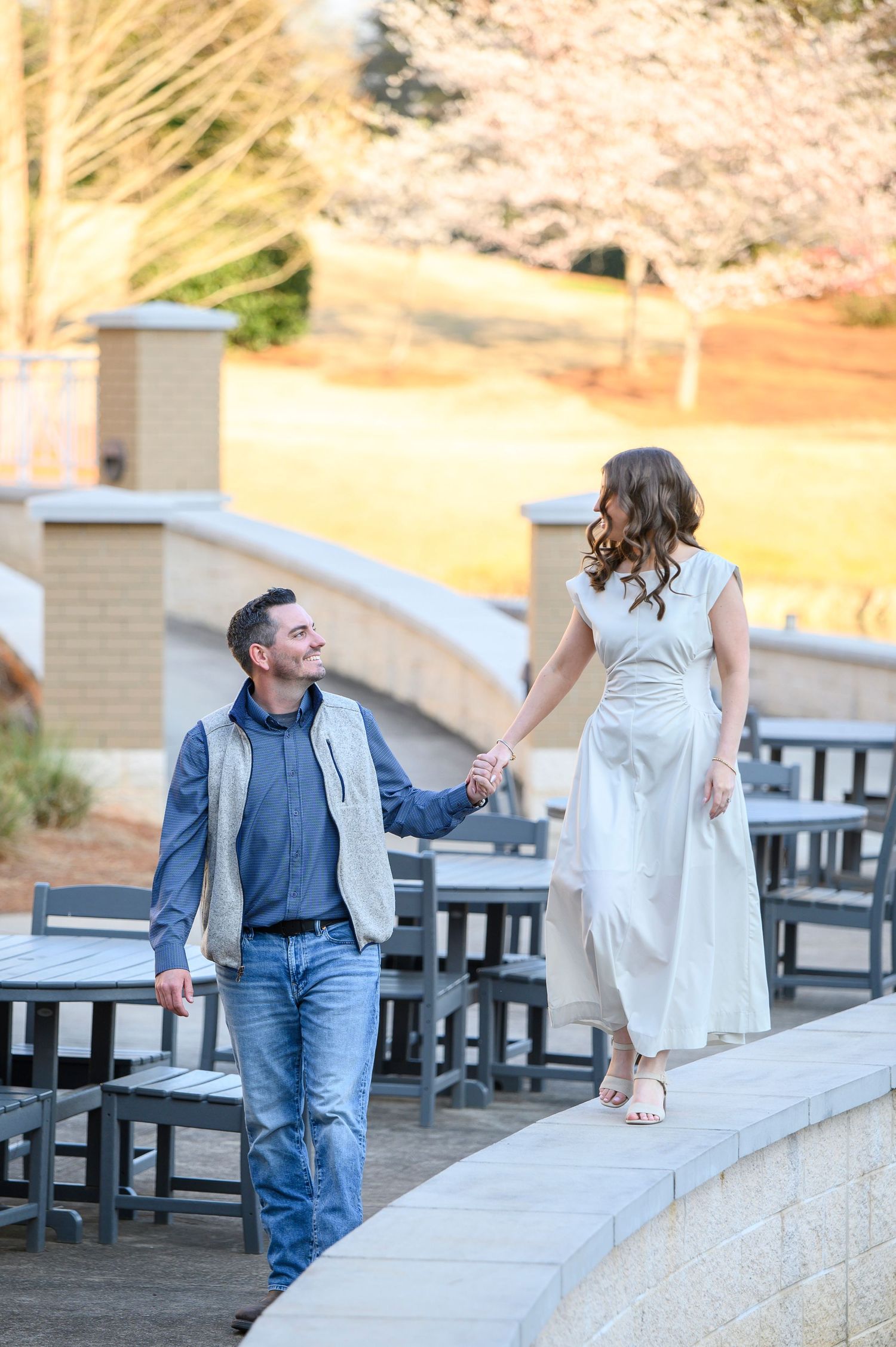 A couple in casual attire sits on a concrete wall outdoors near dining tables during a sunny afternoon.
