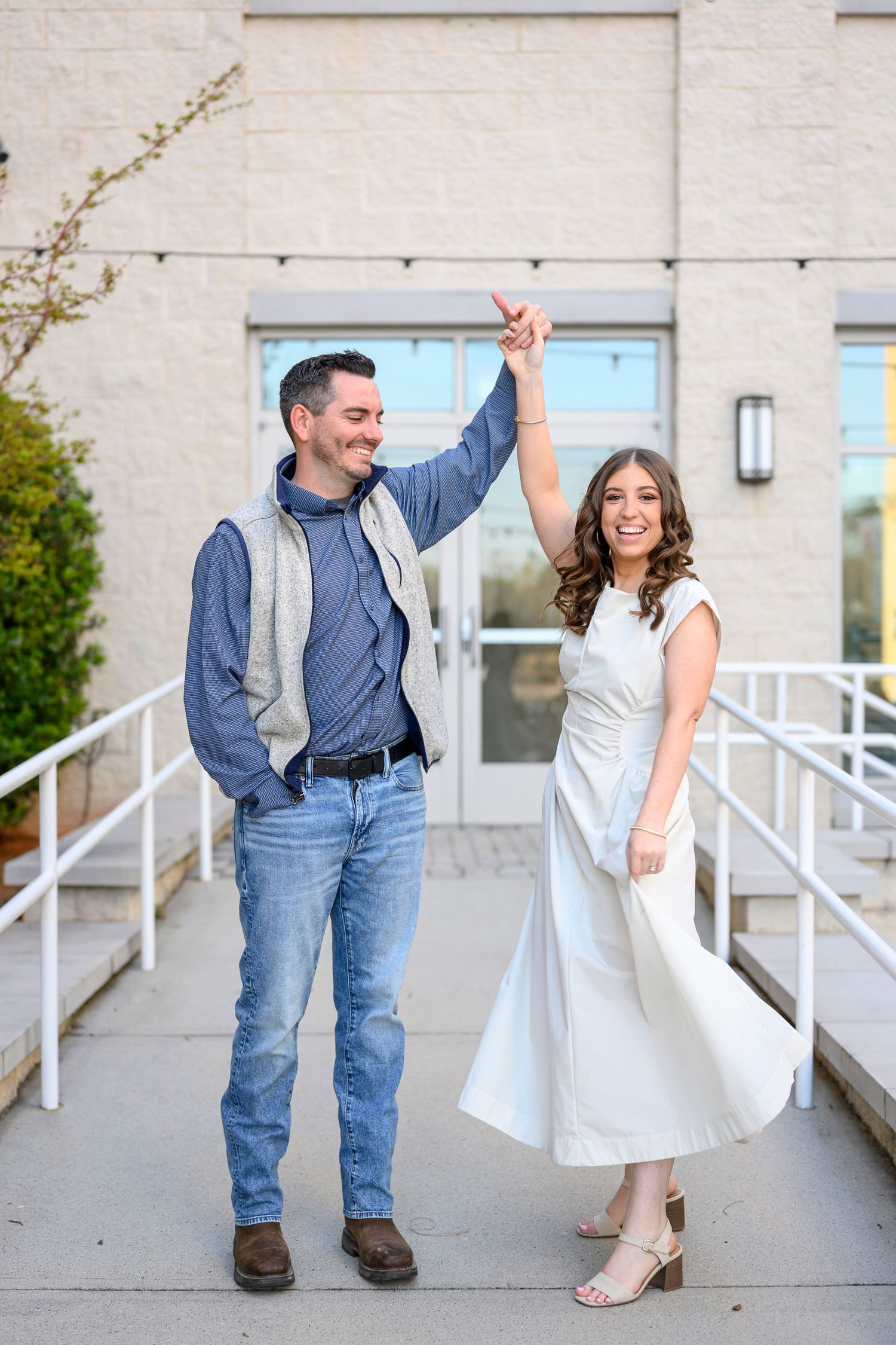 A couple in casual attire dances playfully outside a building with white railings and concrete walls.