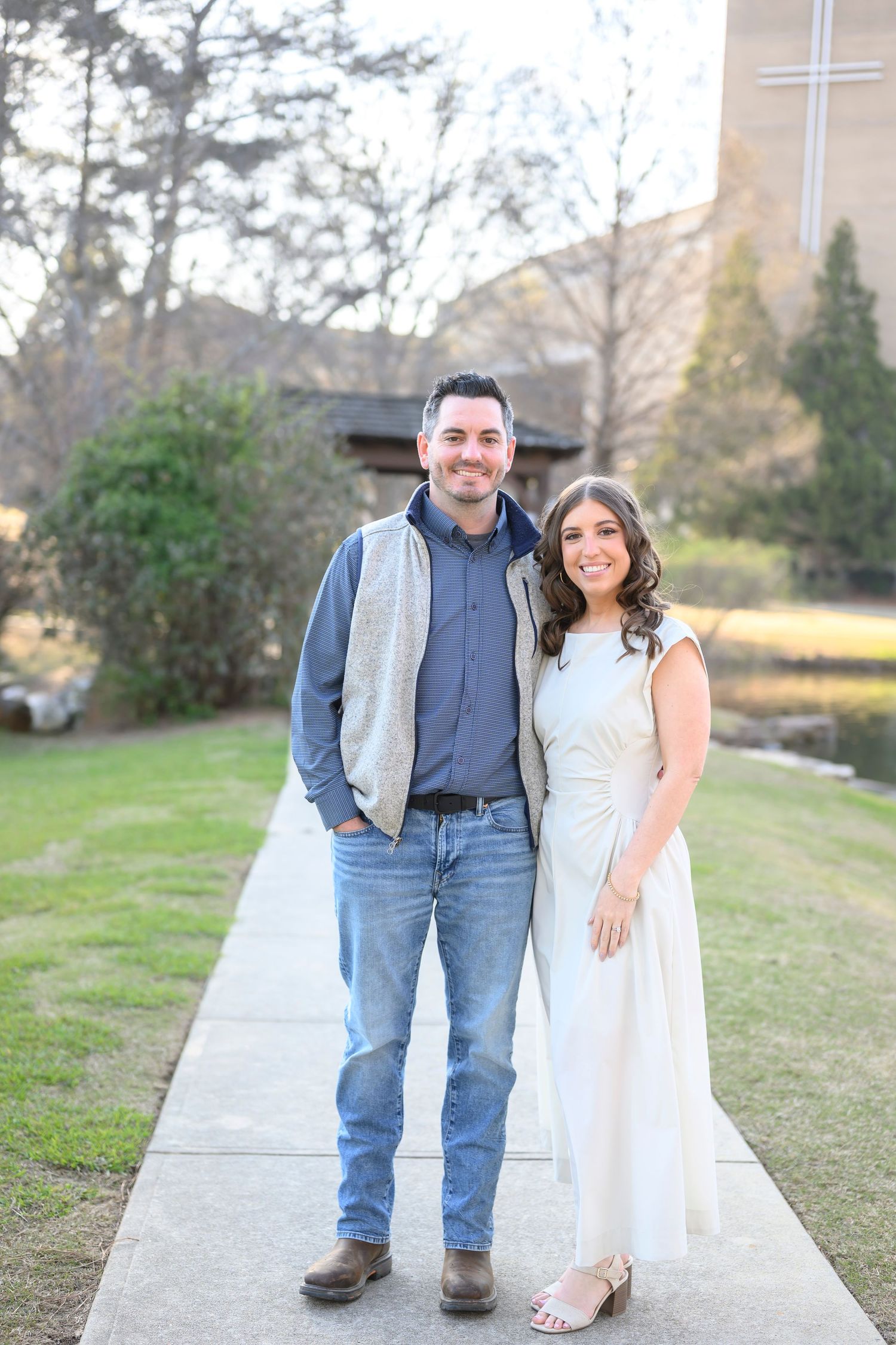 A couple stands together on a paved walkway surrounded by grass and trees on a sunny day.