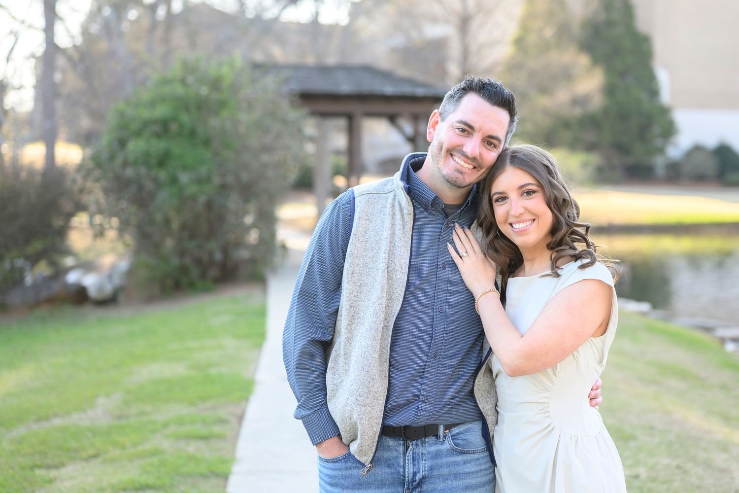 A couple stands close together outdoors on a sunny day with green grass and trees in the background.