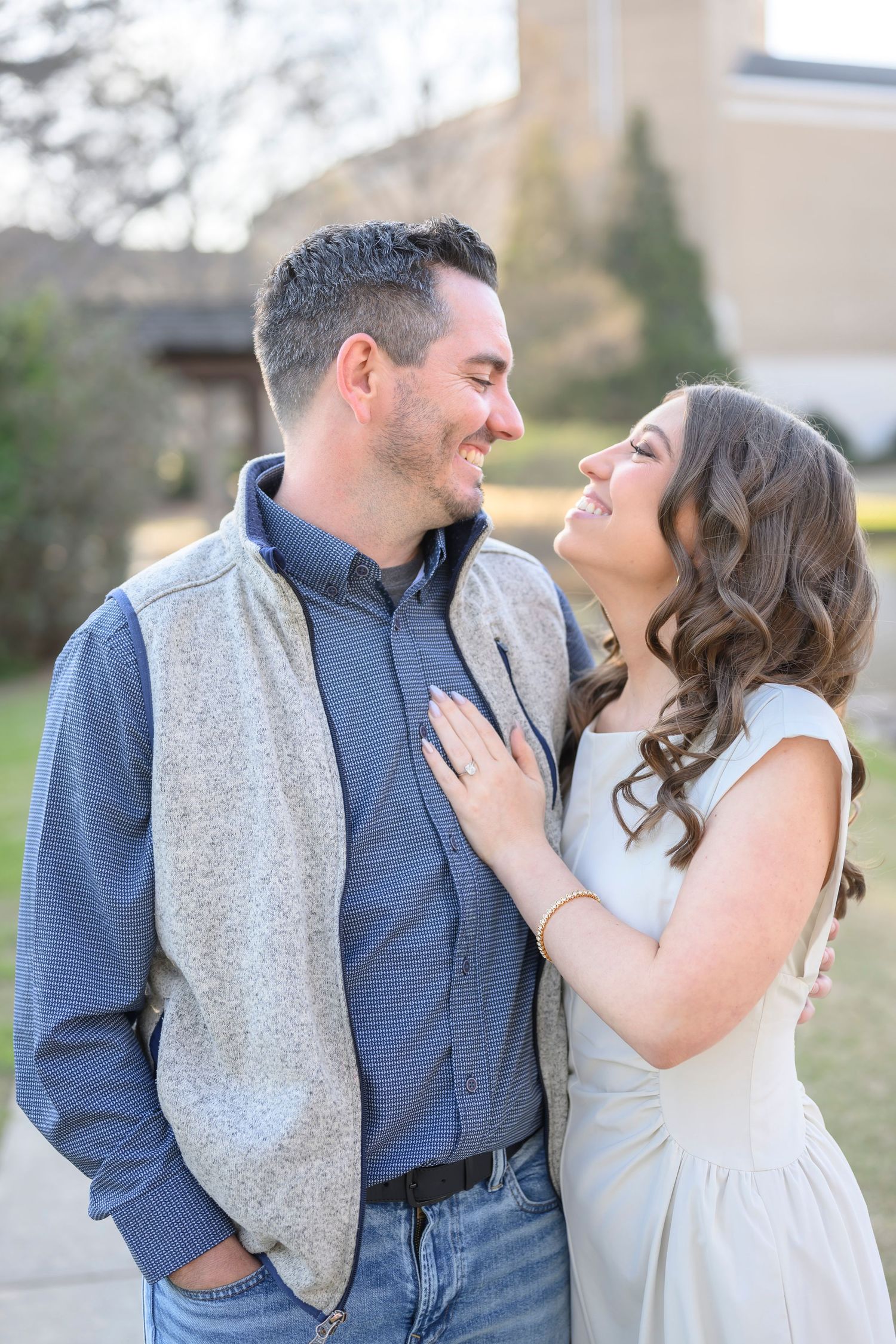 A couple shares a romantic moment outdoors, with the woman in a light dress and man wearing a vest and jeans.
