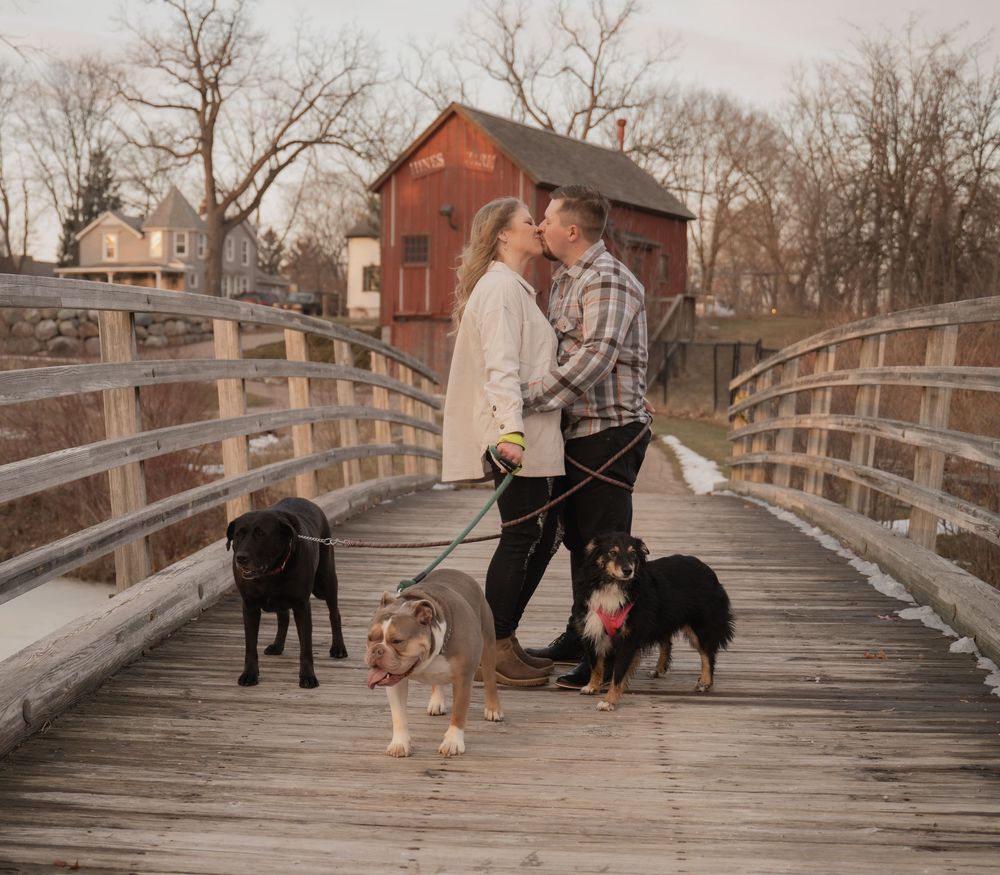 A couple stands on a wooden bridge with three dogs while a red barn sits in the background on an autumn day.