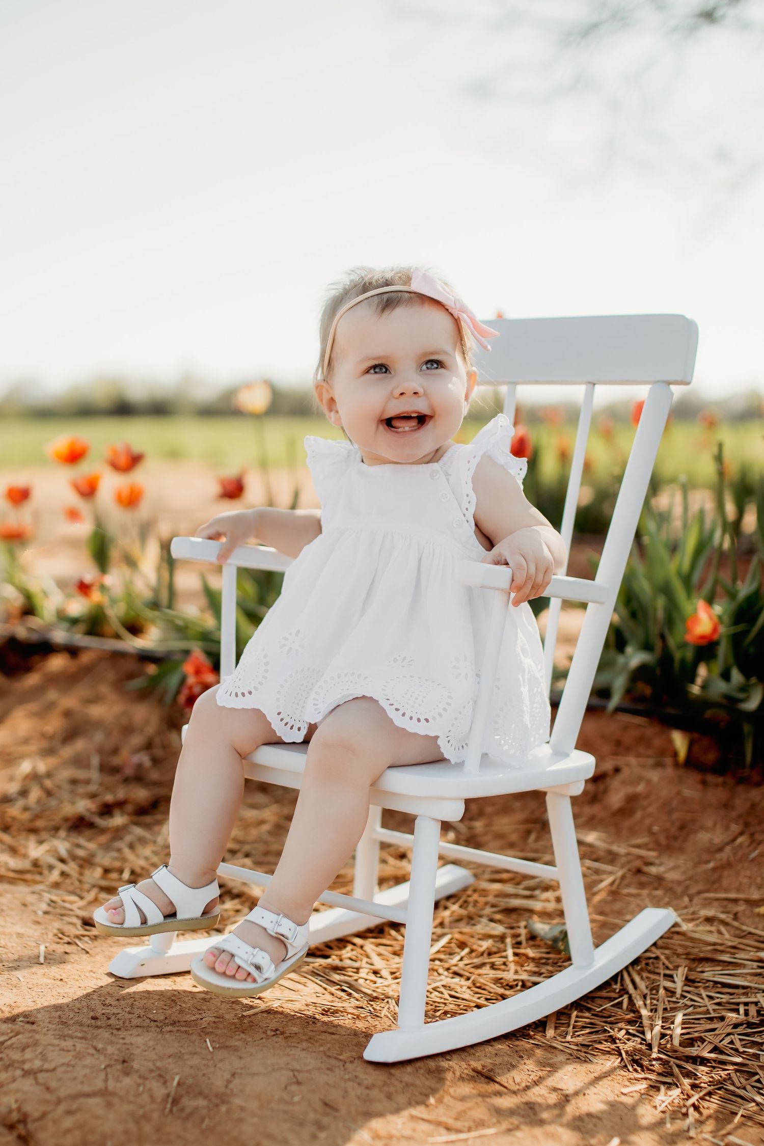 A baby in a white dress sits on a white rocking chair outdoors with orange flowers blooming in the background.