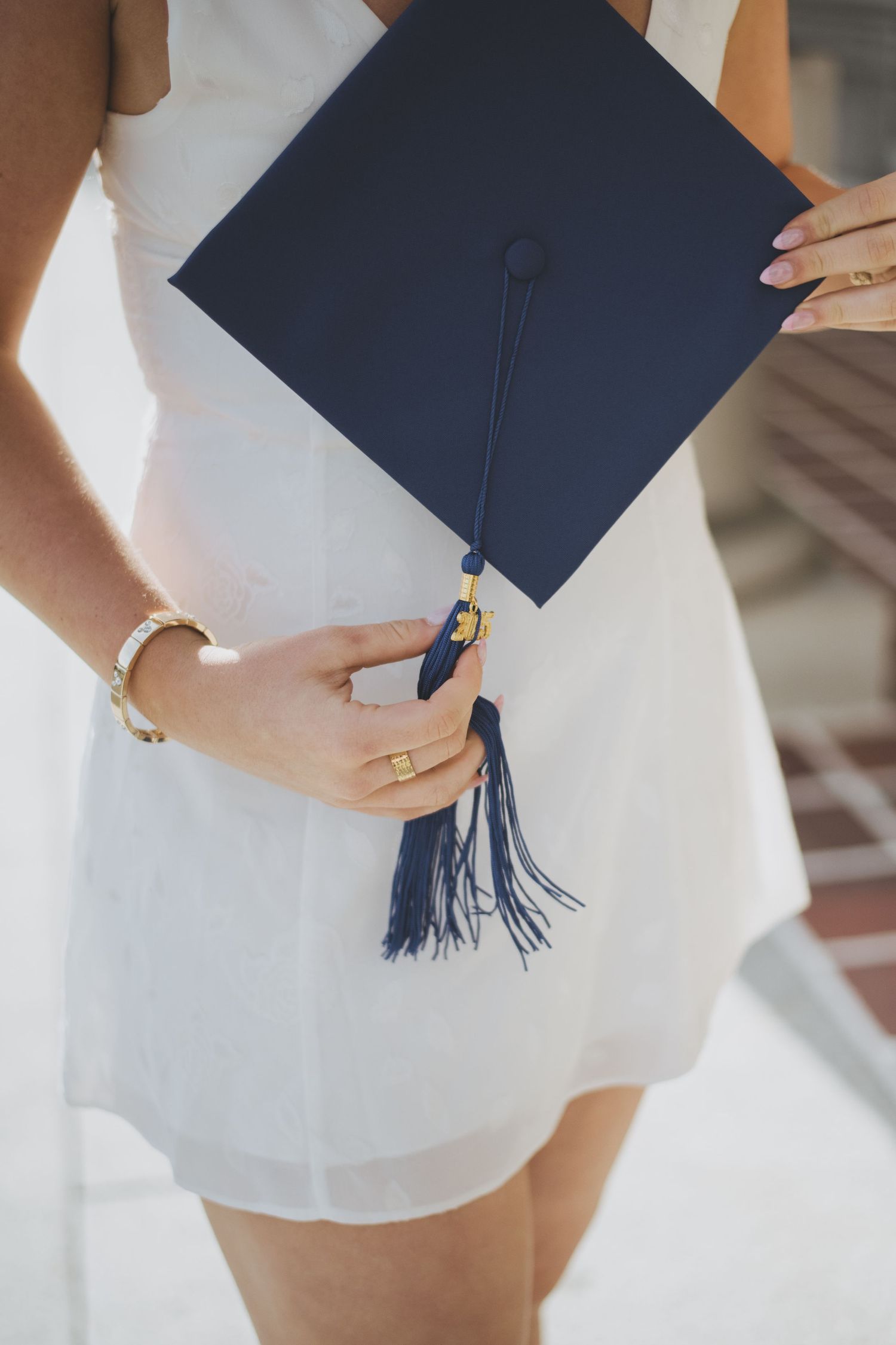 A graduate holds their navy blue mortarboard graduation cap while wearing a white dress and jewelry.