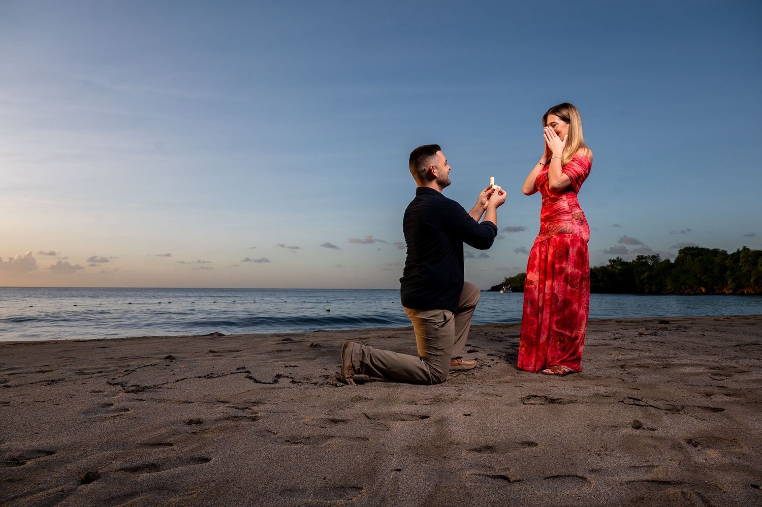 A couple in a red dress and black shirt pose together on a sandy beach at sunset during a photoshoot.