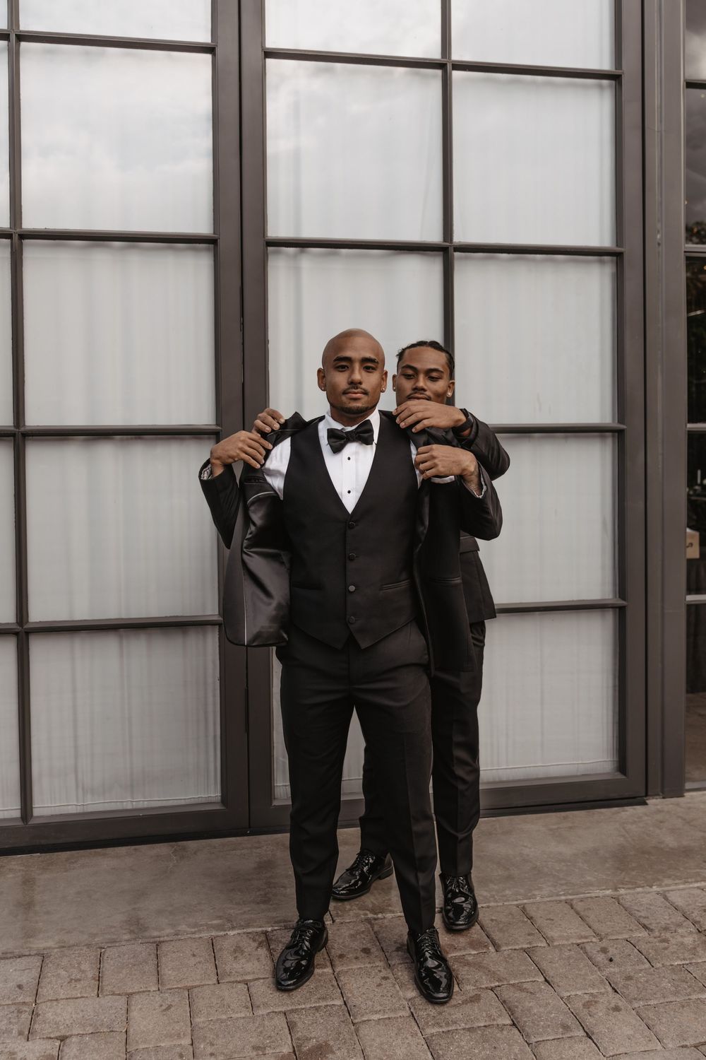 Two people in formal black tuxedos pose together against a modern glass wall backdrop.