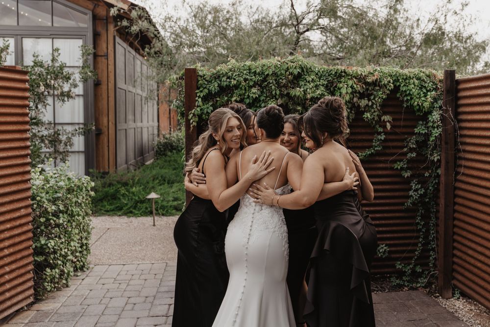 Three women in black dresses embrace a bride wearing a fitted white wedding gown in an outdoor garden setting.