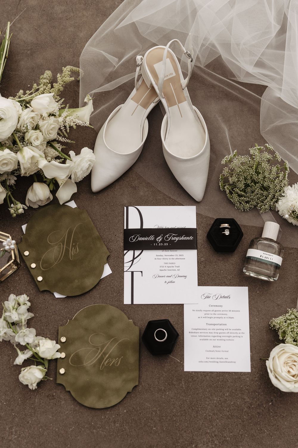 Wedding details flatlay featuring white bridal shoes, invitation cards, rings, and white roses on a dark background.