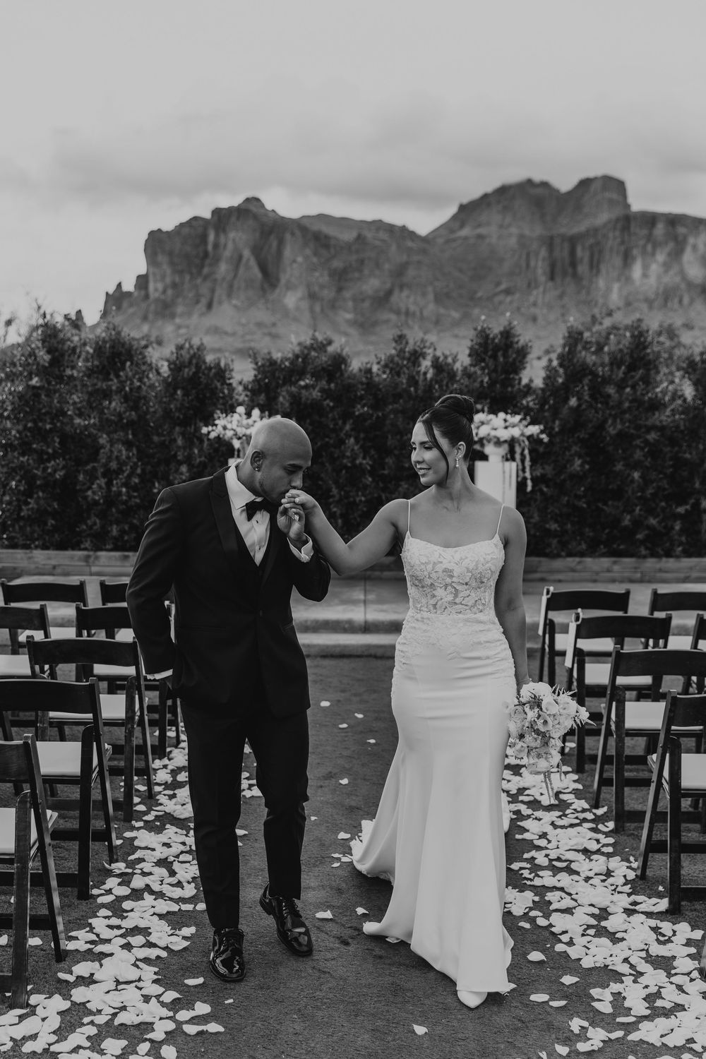 Black and white wedding ceremony sequence showing couple walking down petal-lined aisle with mountain backdrop.