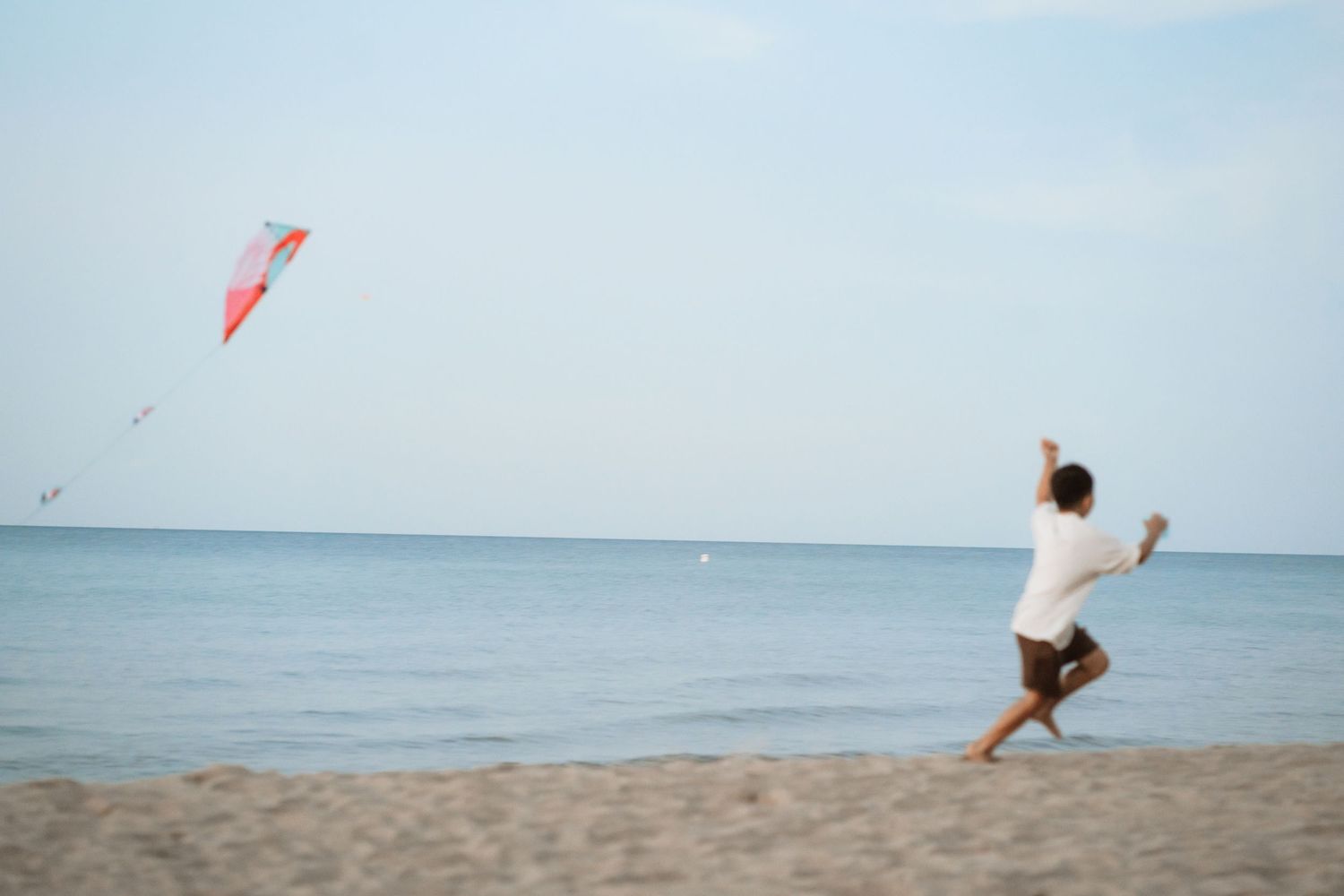A person flies a red kite on a sandy beach against a bright blue ocean and sky backdrop.