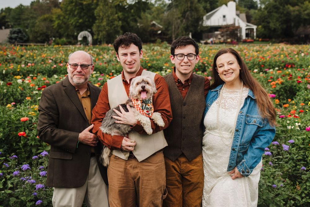 Family of four poses with their dressed-up dog in a vibrant flower field, smiling for a portrait.