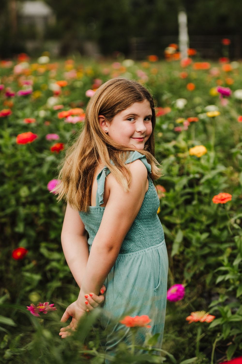 A young girl in a teal dress smiles over her shoulder while standing in a vibrant field of colorful zinnia flowers.