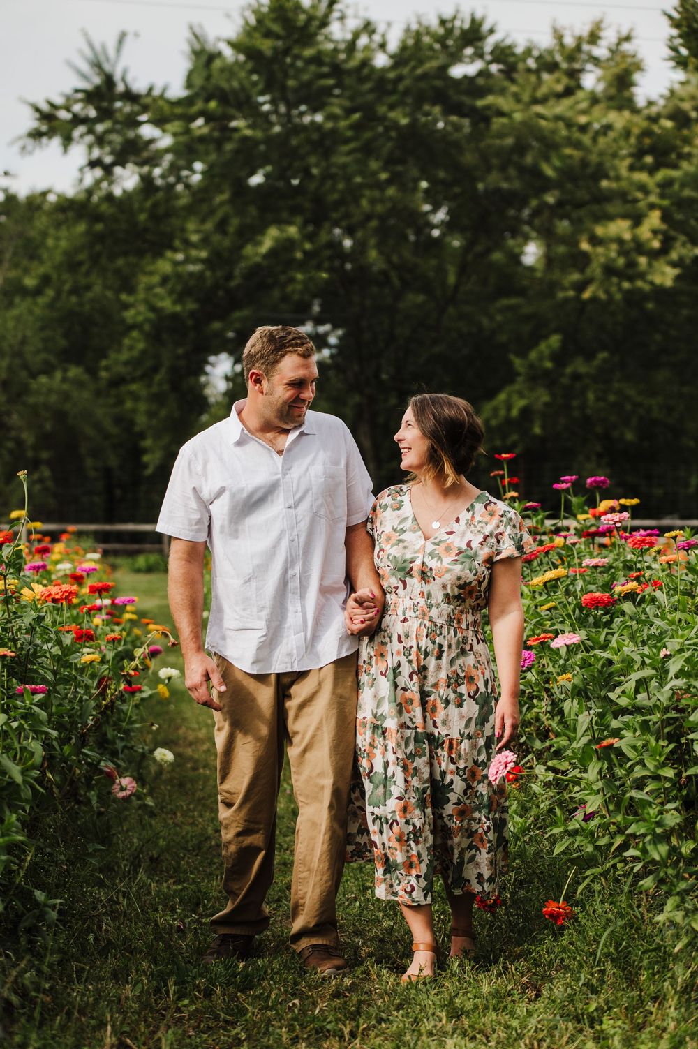 A couple walks hand-in-hand through a vibrant flower garden, smiling at each other, surrounded by colorful zinnias and lush green trees.