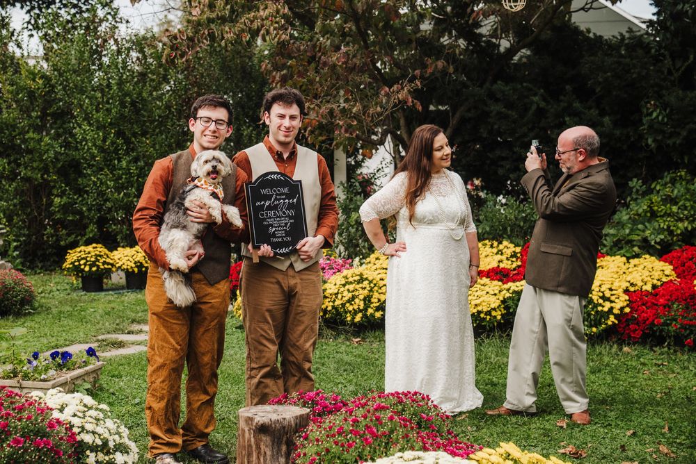 Outdoor wedding ceremony with groom holding dog, bride in white dress, and guests among colorful flower gardens.