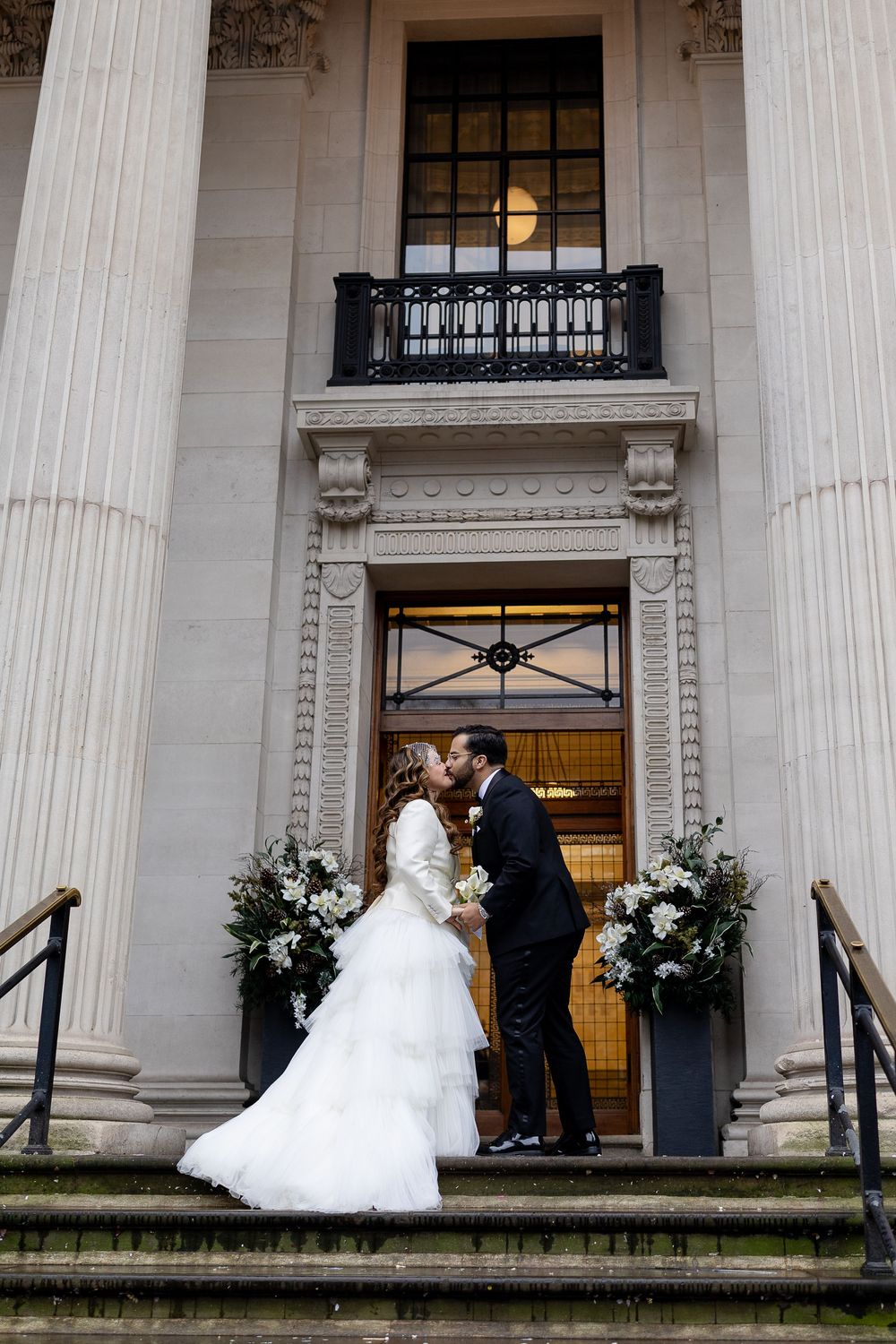 A wedding ceremony takes place on the steps of a grand neoclassical building with white columns.