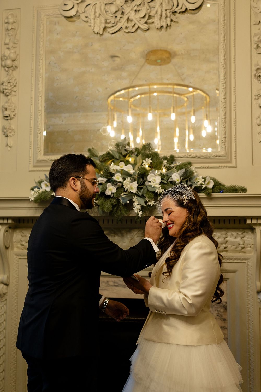 A romantic wedding couple shares an intimate moment in an elegant room with ornate white decor and chandeliers.