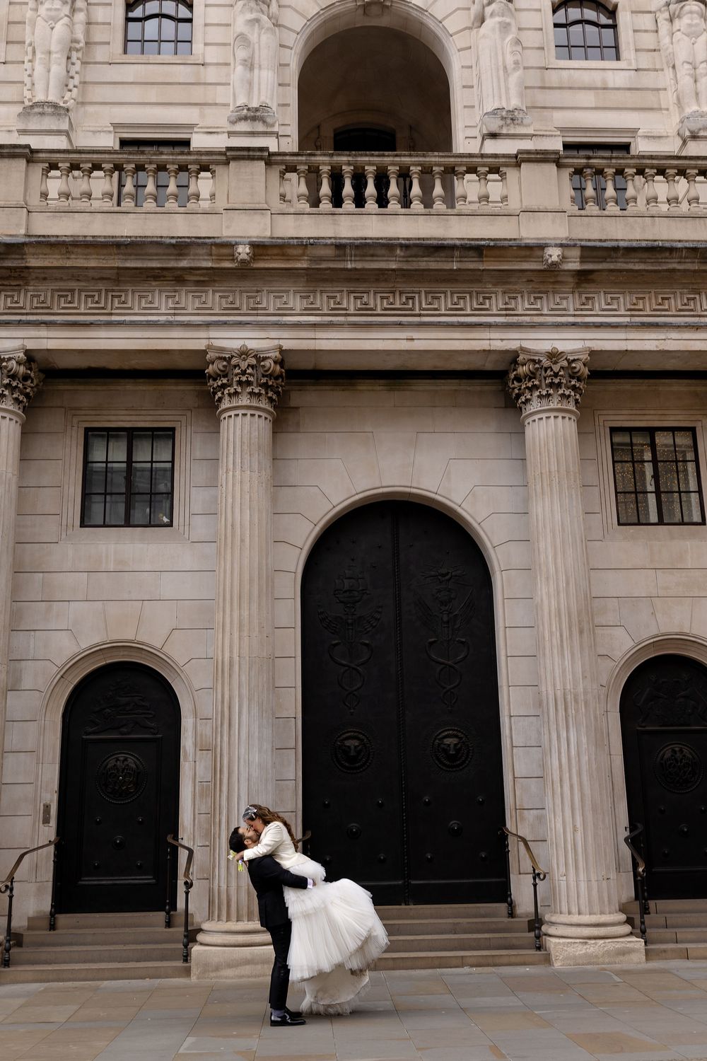 Elegant wedding couple poses in front of ornate stone building with classical architecture.