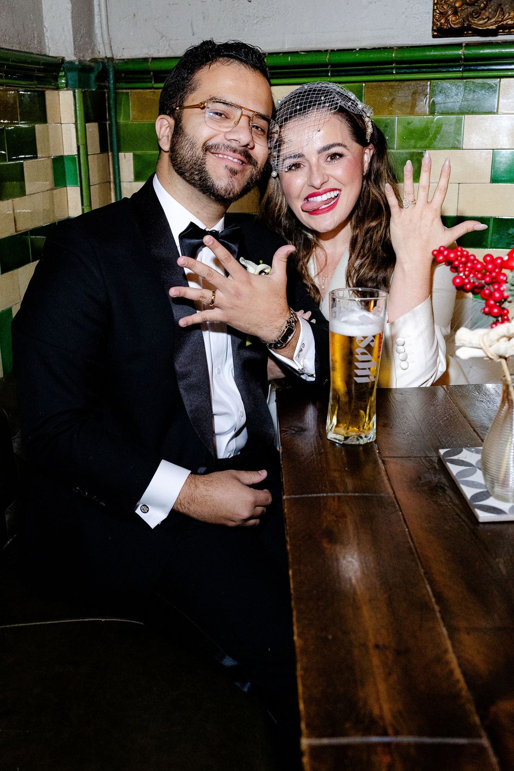 A couple in formal attire sits at a wooden table with beers celebrating a special occasion at a pub.