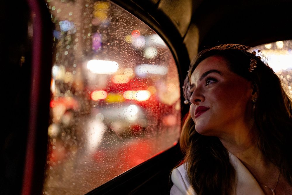 Person gazing out rainy window of car at night with blurred city lights in background.