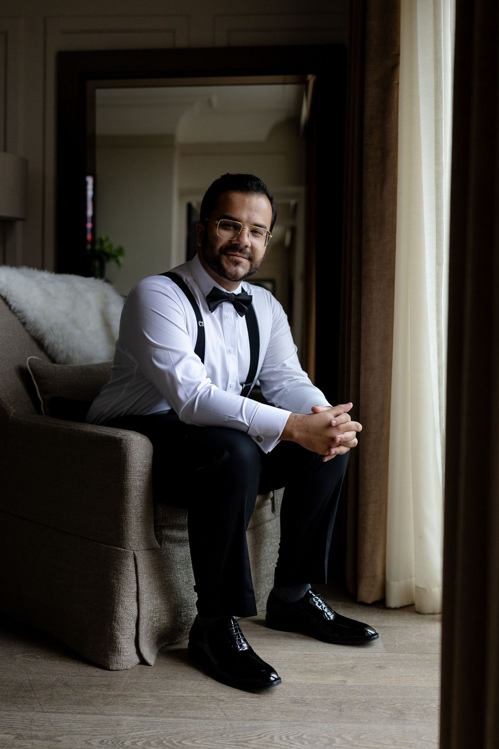 Groom in formal attire with bow tie and suspenders sits pensively in a hotel room before wedding ceremony.