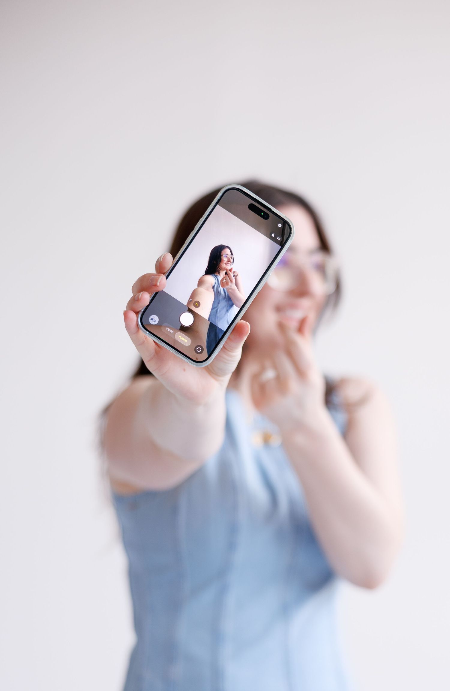 Someone in a light blue top holds up a smartphone displaying a selfie photo on its screen against a white background.