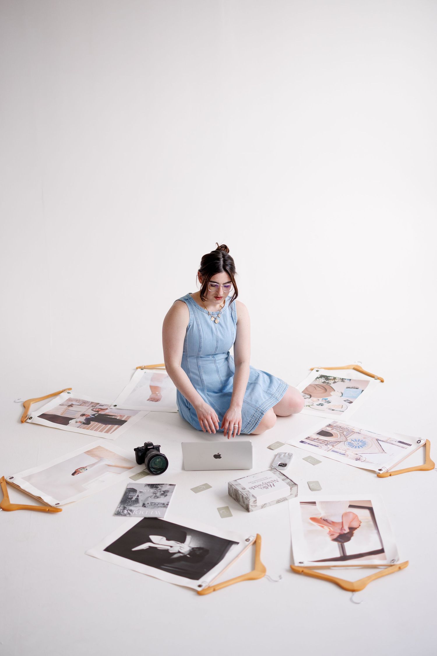 Creative workspace with photographer sitting on floor surrounded by printed photos and laptop against white background.