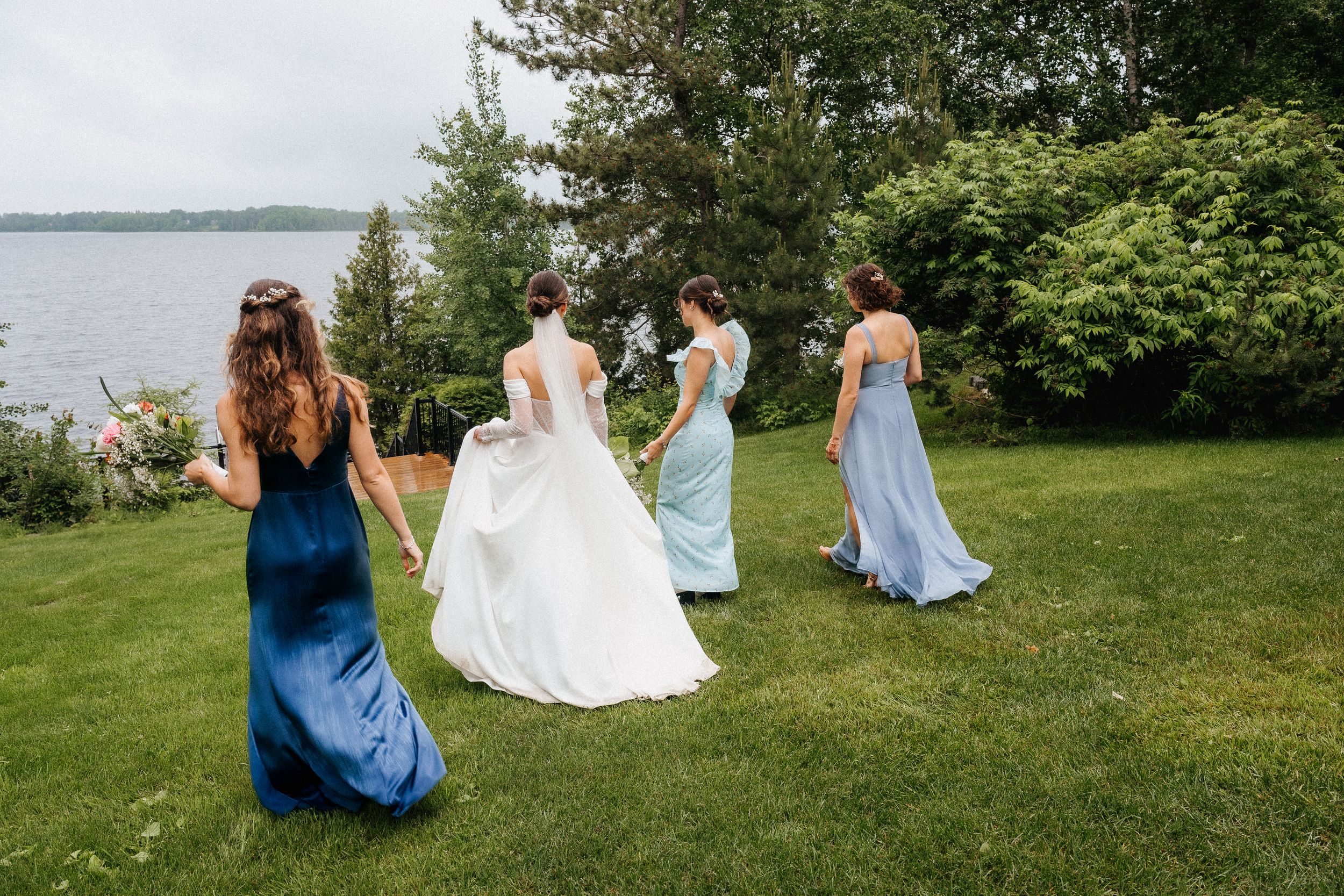 Bridesmaids in flowing blue dresses walk with bride in white gown across green lawn overlooking waterfront.