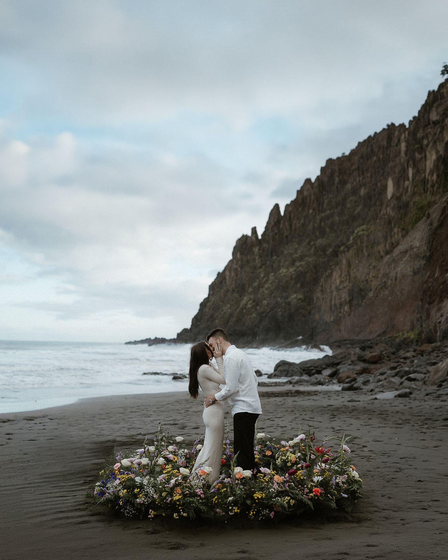 A romantic couple embraces on a black sand beach with dramatic volcanic rock formations in the background.