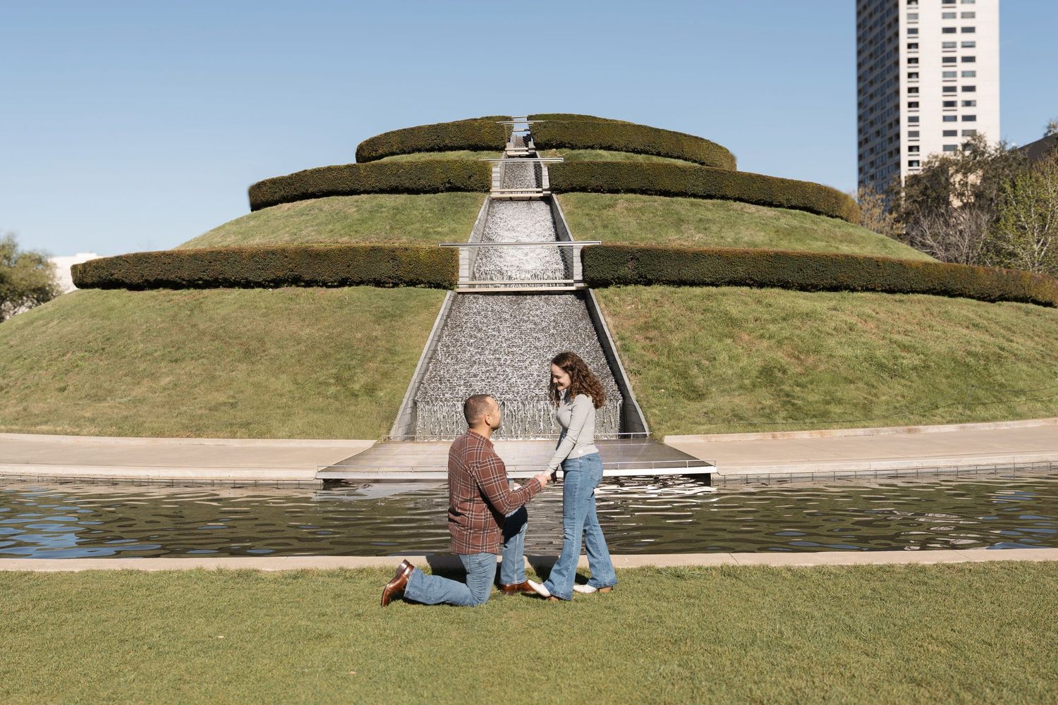 Panoramic view of grassy hills with modern buildings and monuments in an urban park setting.