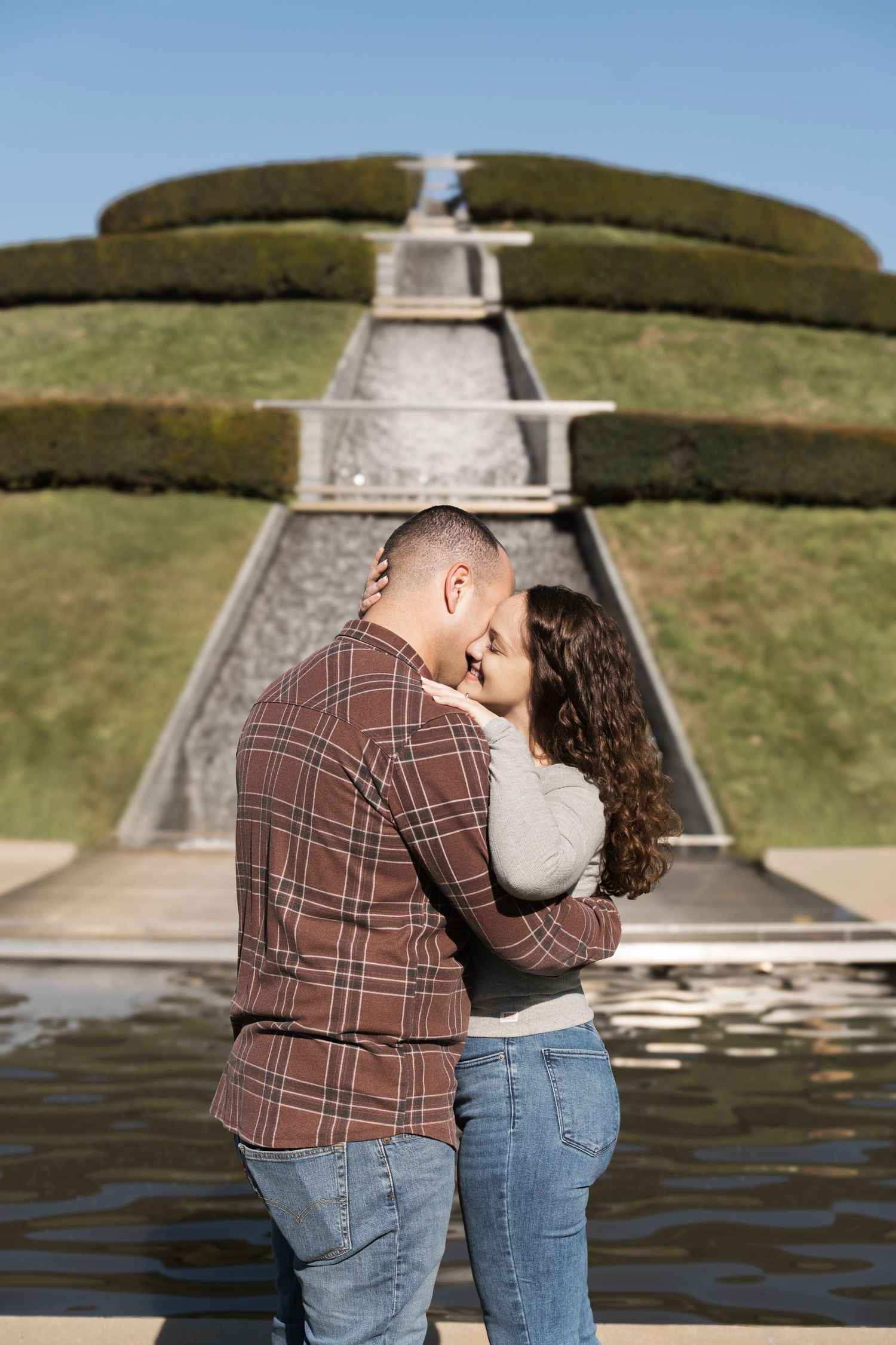 A couple embraces in a romantic moment by a waterfront with rolling green hills and stone steps in the background.