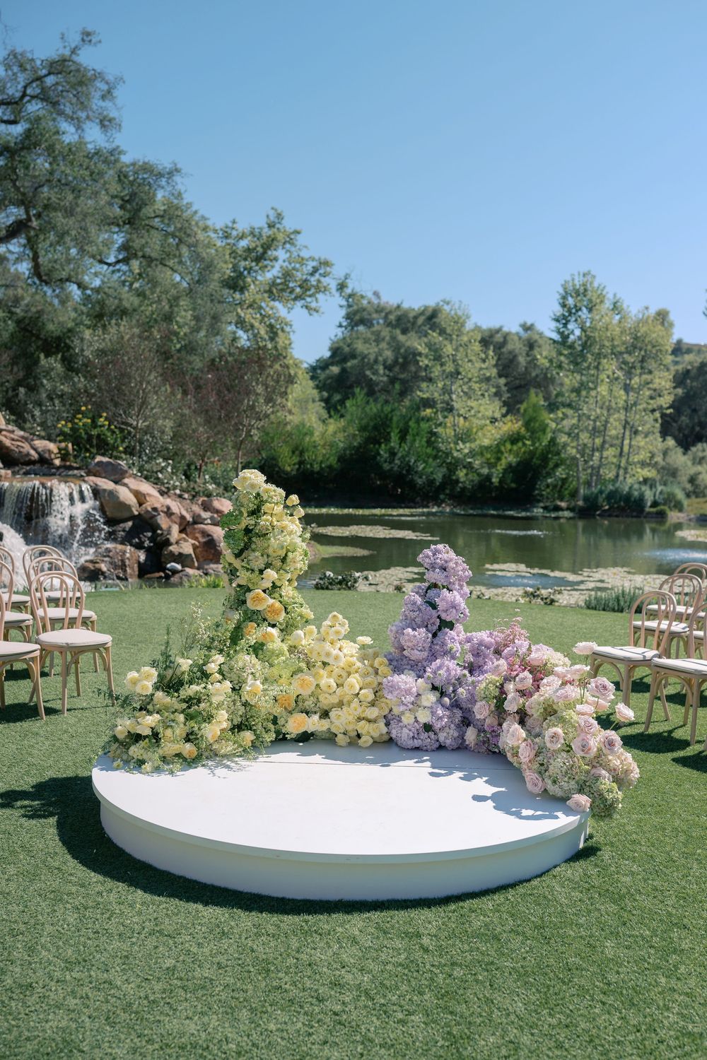 Outdoor wedding ceremony setup with curved white platform and pastel floral arrangements on grassy lawn by pond.