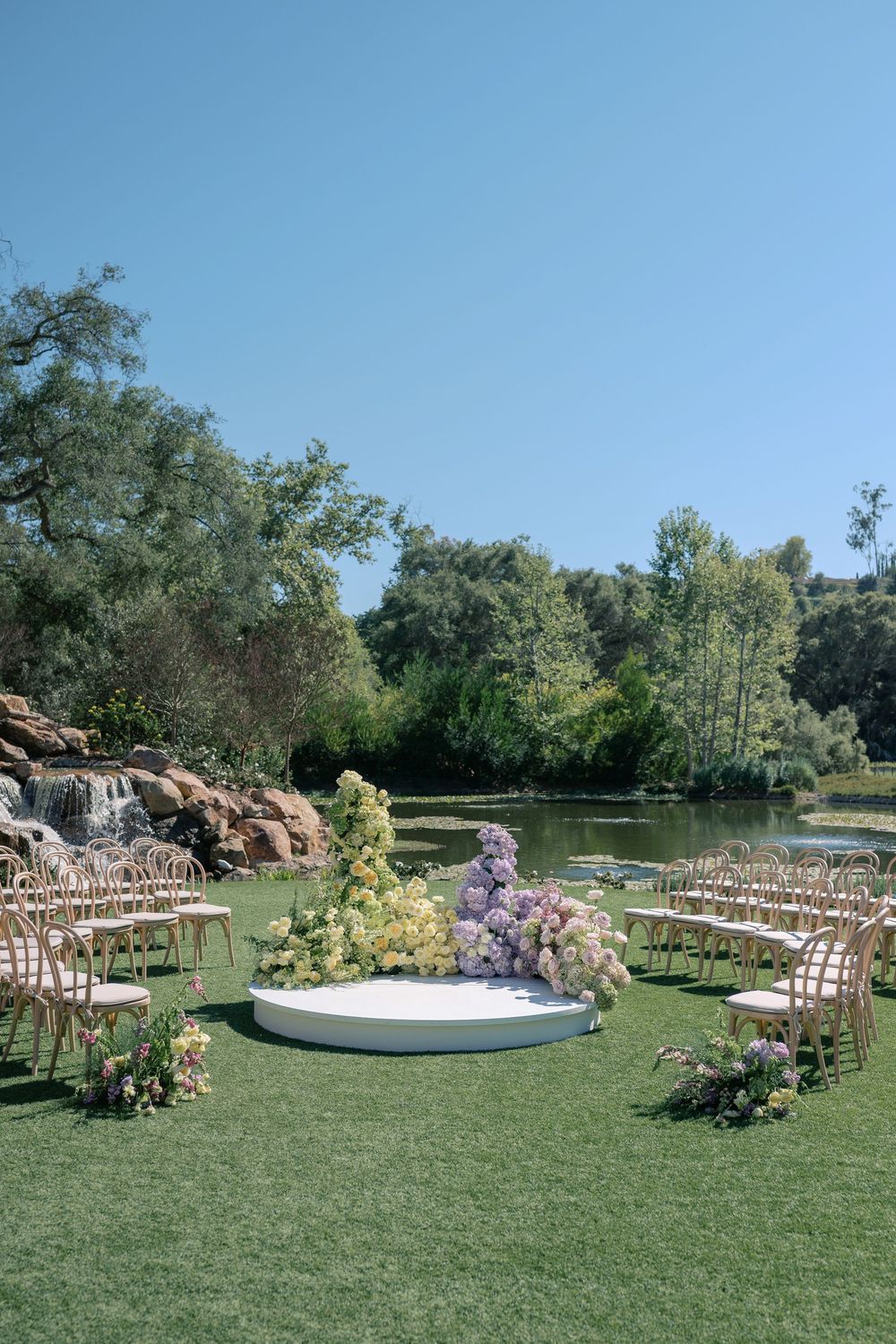 Outdoor wedding ceremony setup with wooden chairs and floral arrangements around a circular platform on a grassy lawn.