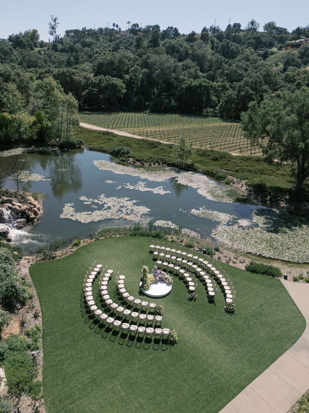 Aerial view of outdoor wedding ceremony setup with chairs arranged in curved rows on a lush green lawn beside a pond.