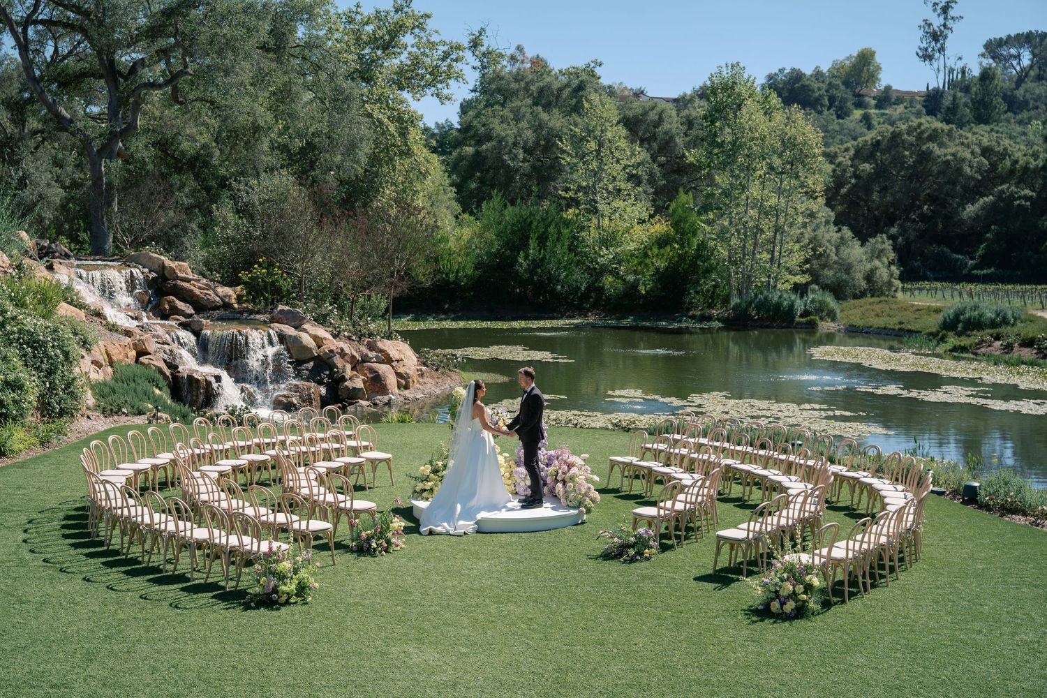 Curved rows of wooden chairs arranged outdoors for a garden wedding ceremony beside a tranquil pond.