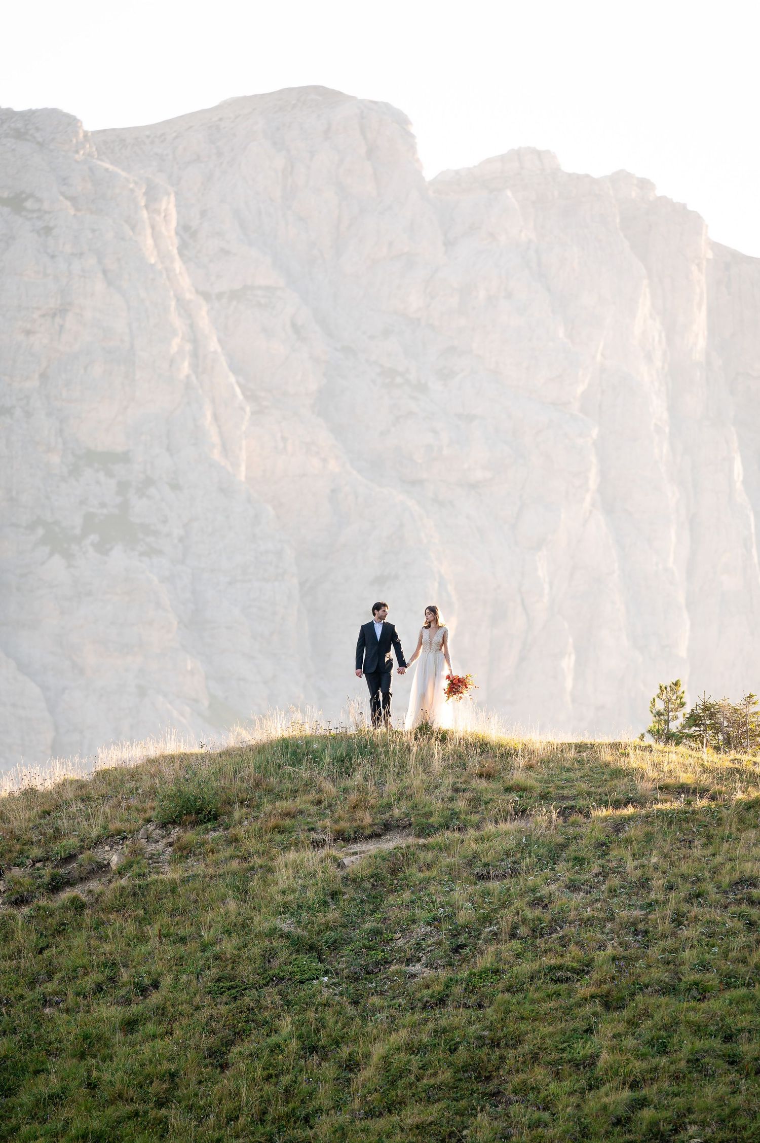 Two silhouetted figures walk along a grassy hilltop ridge against dramatic mountain cliffs in golden evening light.