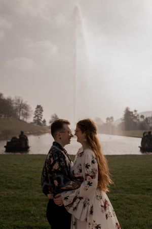 A romantic couple embraces near a fountain in a misty park setting during golden hour.