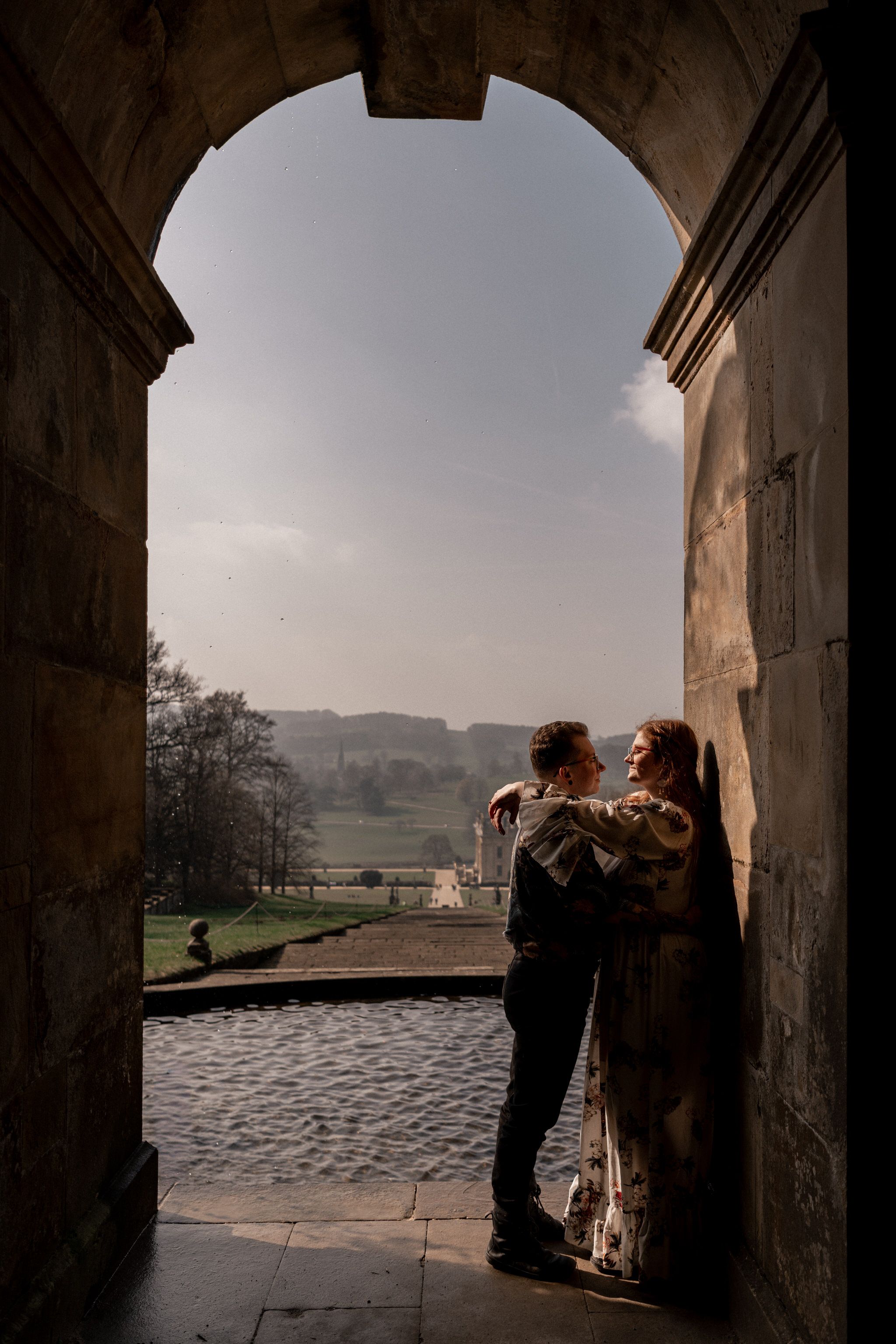 A couple stands silhouetted in a stone archway overlooking a misty countryside landscape.
