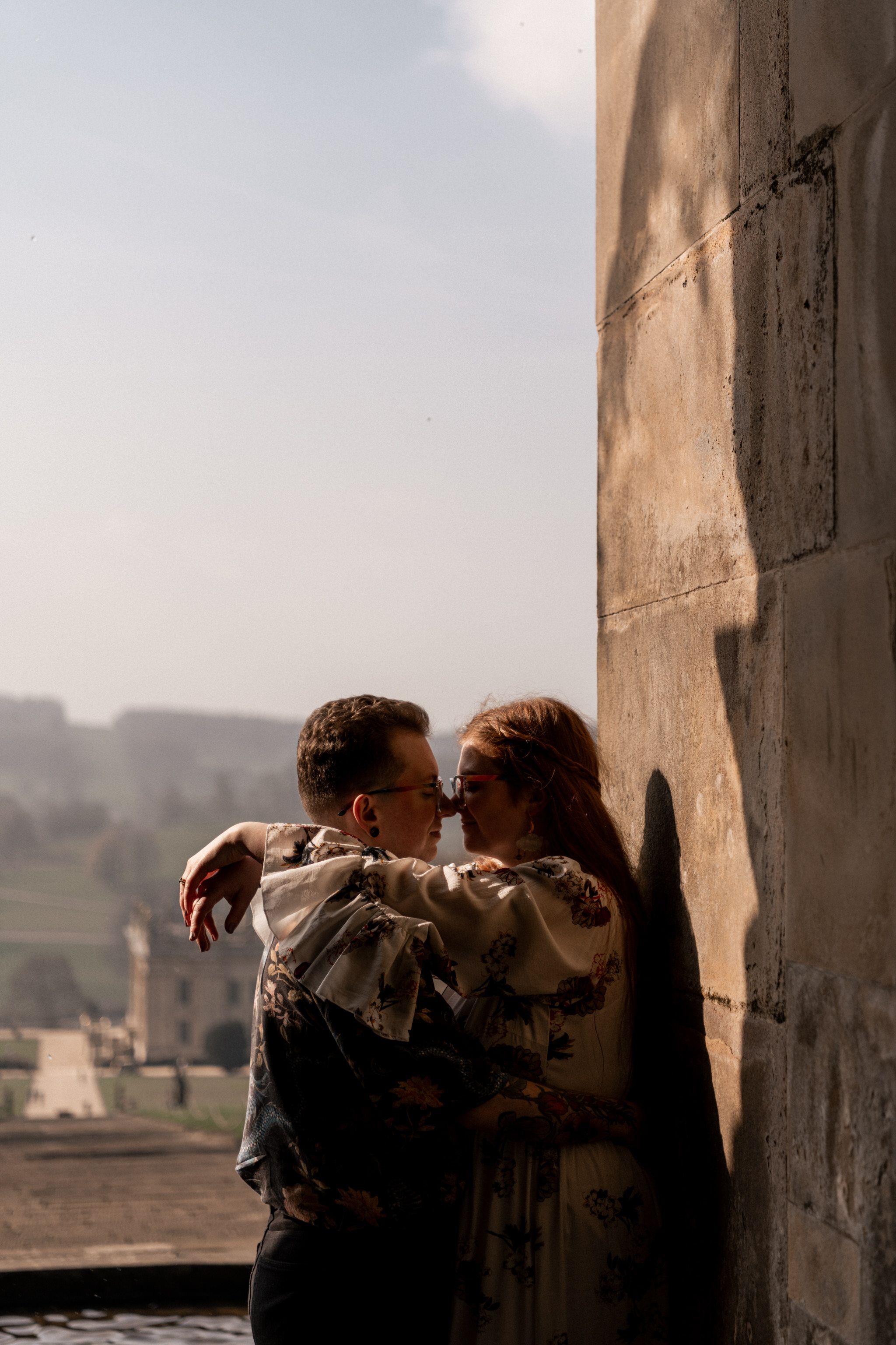 A romantic couple shares an intimate moment in an archway overlooking a distant landscape at sunset.