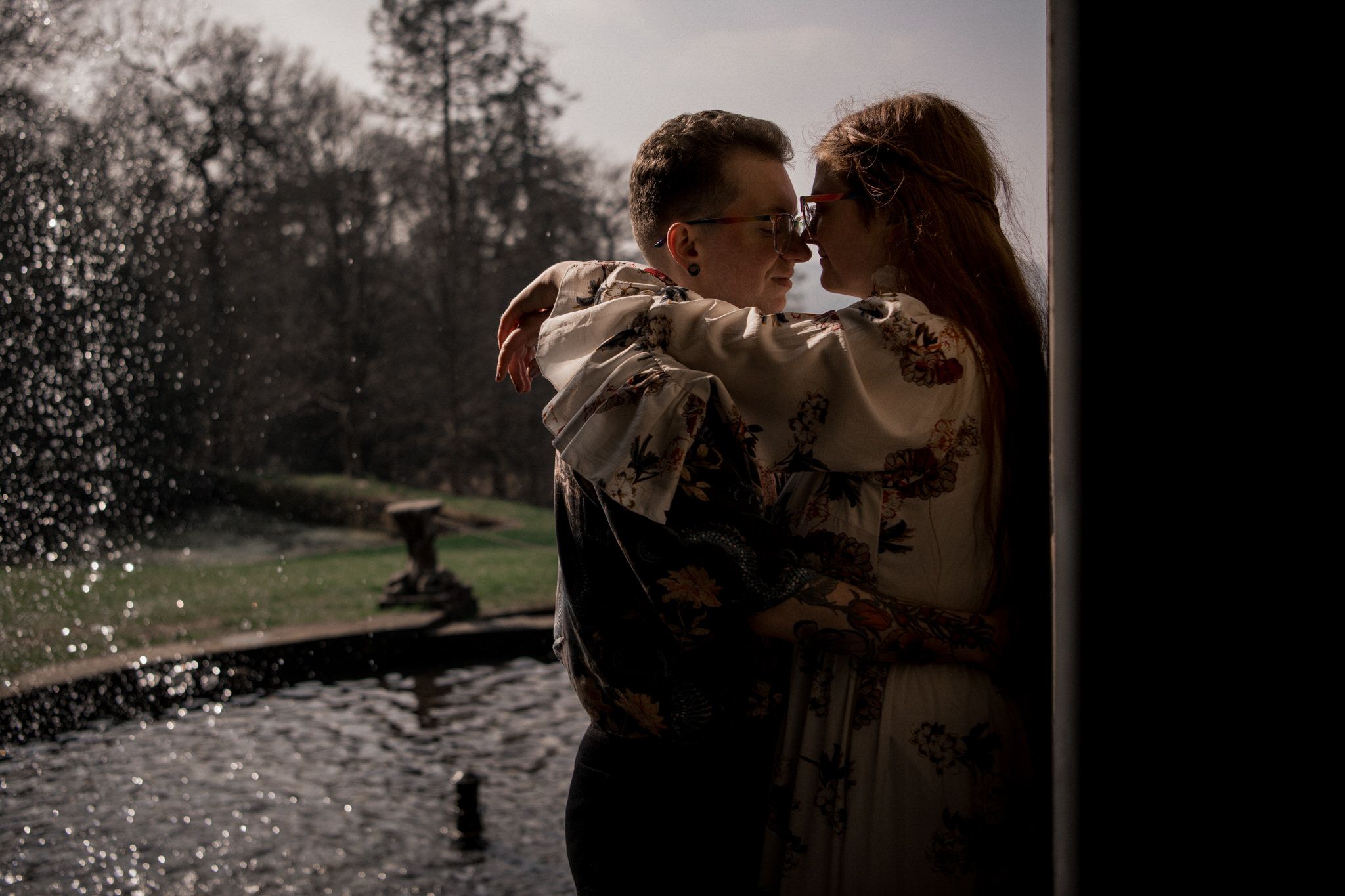 A romantic couple embraces in silhouette near a fountain on a misty day in a park setting.
