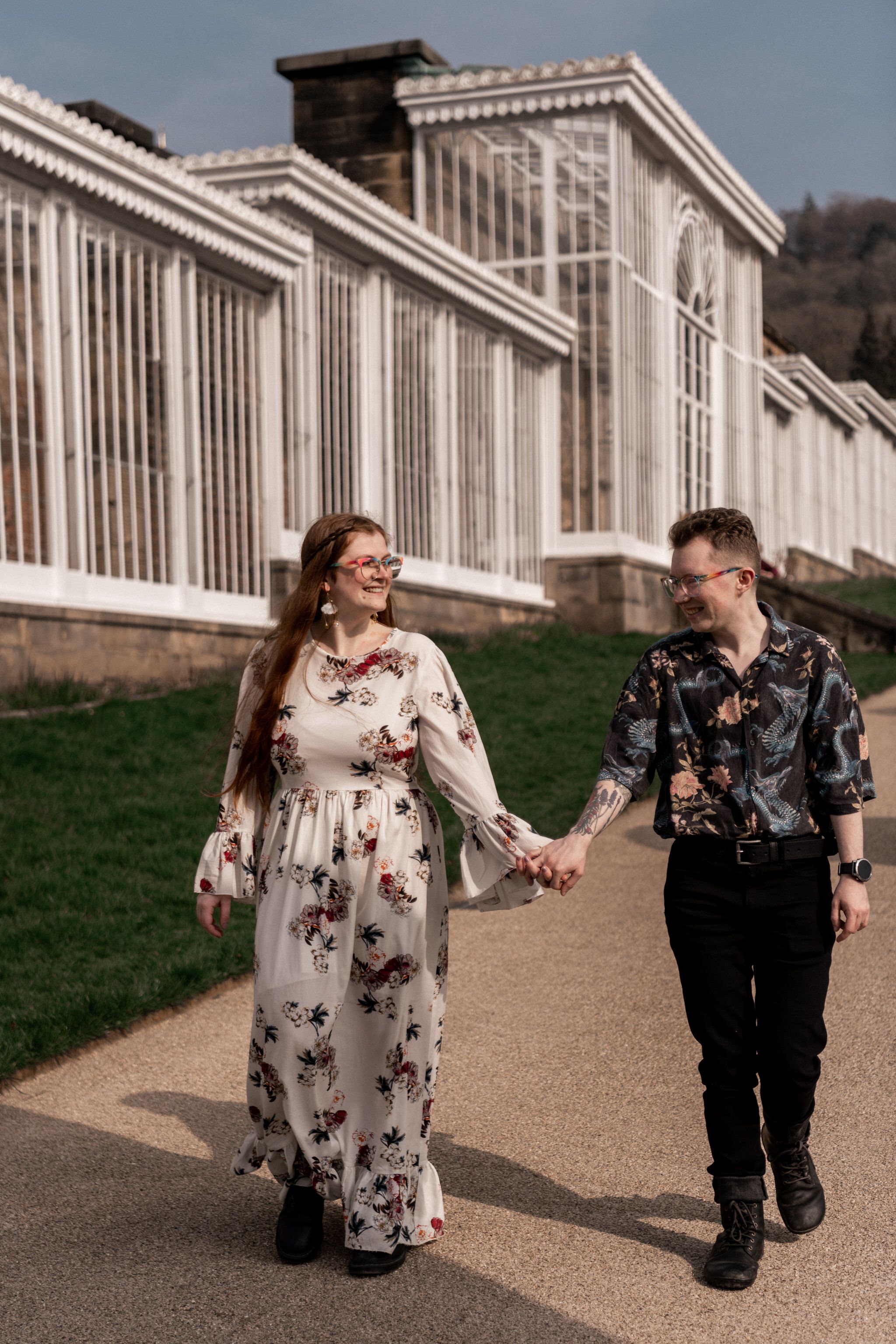 A couple walks hand in hand along a path outside a grand conservatory with white columns and glass panels.
