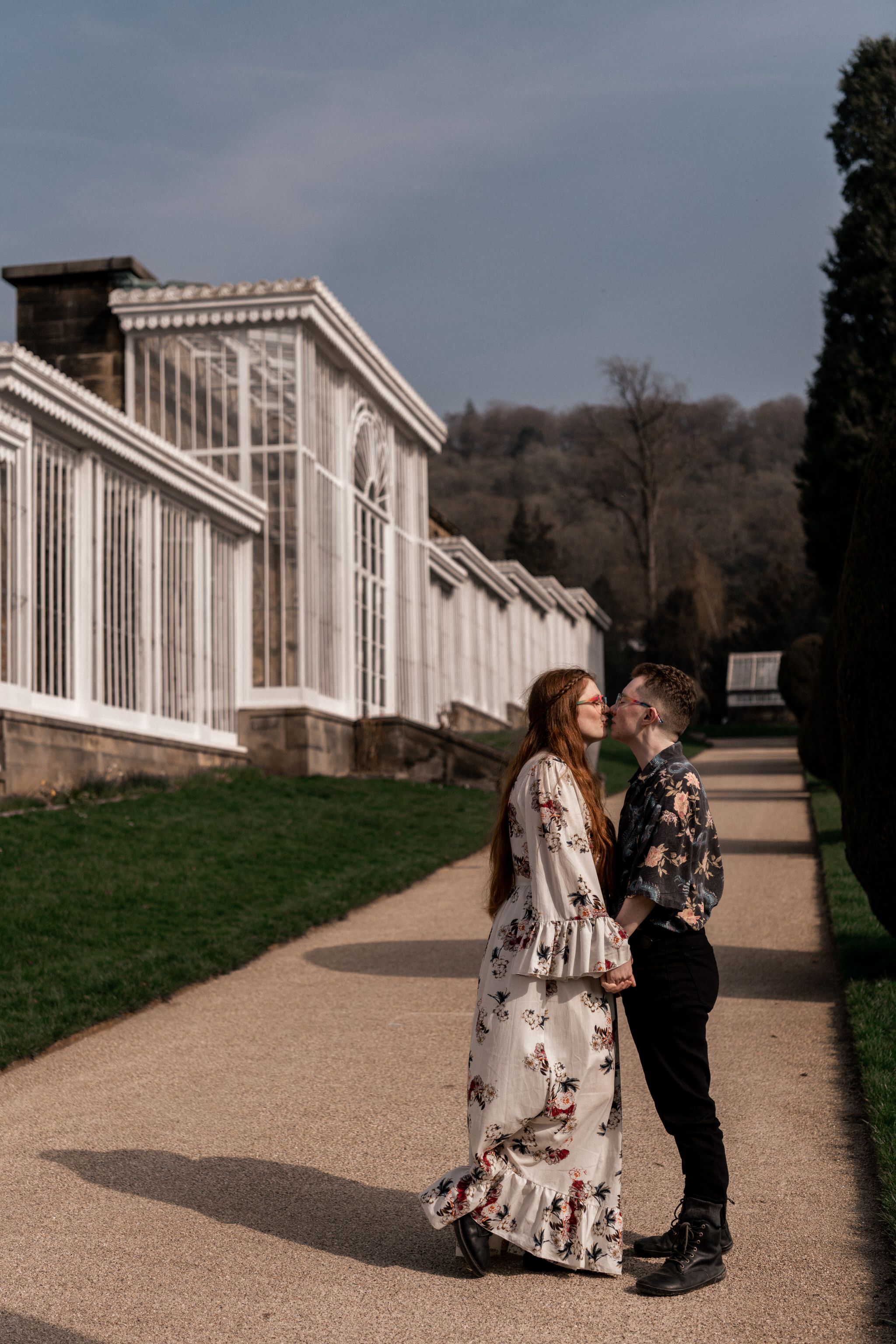 A couple stands together on a path outside a historic Victorian greenhouse with white columns and glass panels.