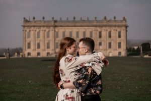 A couple poses for romantic engagement photos in front of a grand historic mansion with ornate architecture.