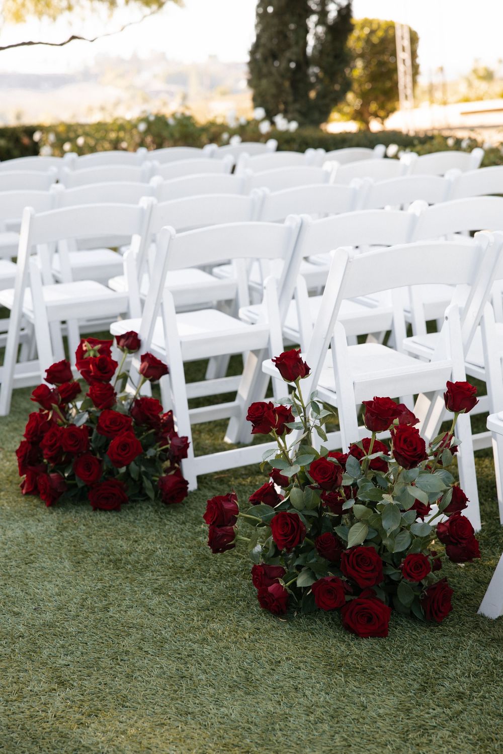 White folding chairs arranged on grass with elegant red rose bouquets lining the aisle at an outdoor wedding ceremony.