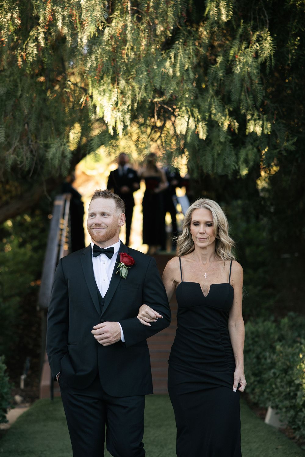 A man in a black tuxedo and woman in a black gown walk together outdoors under lush green trees.