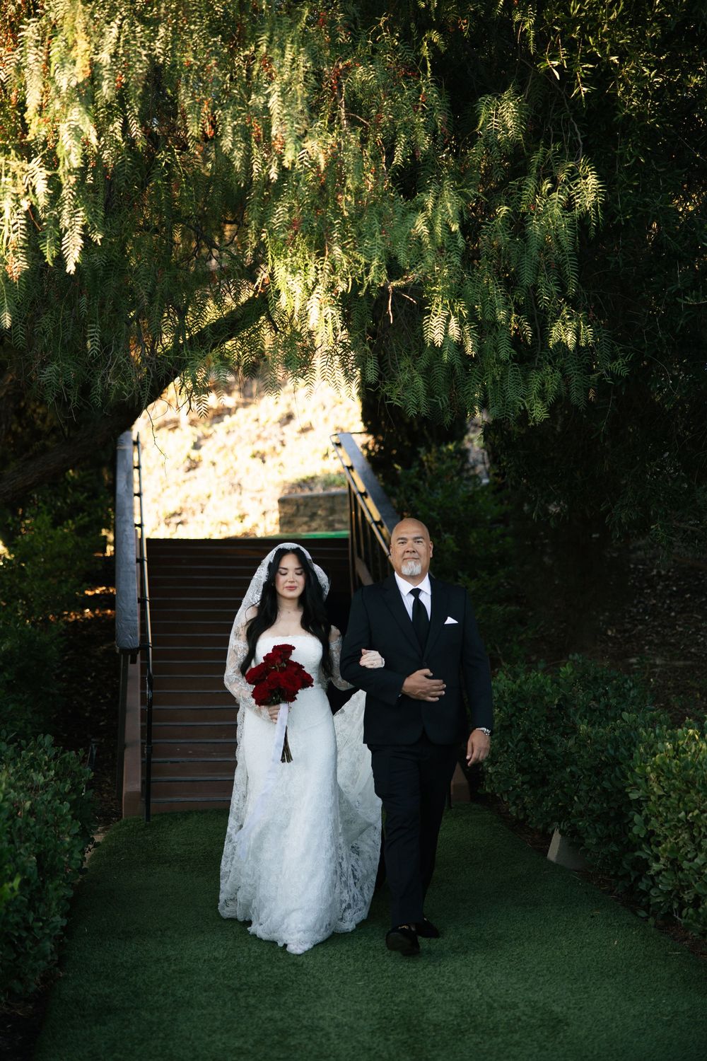 Bride in white lace gown holding red roses walks with suited man down garden path beneath weeping trees.