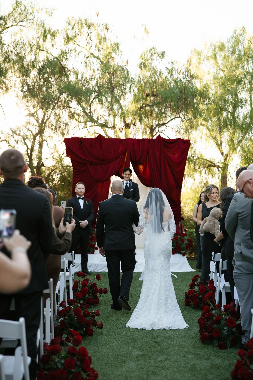 Bride walks down aisle lined with red roses toward a crimson velvet altar backdrop at an outdoor wedding ceremony.