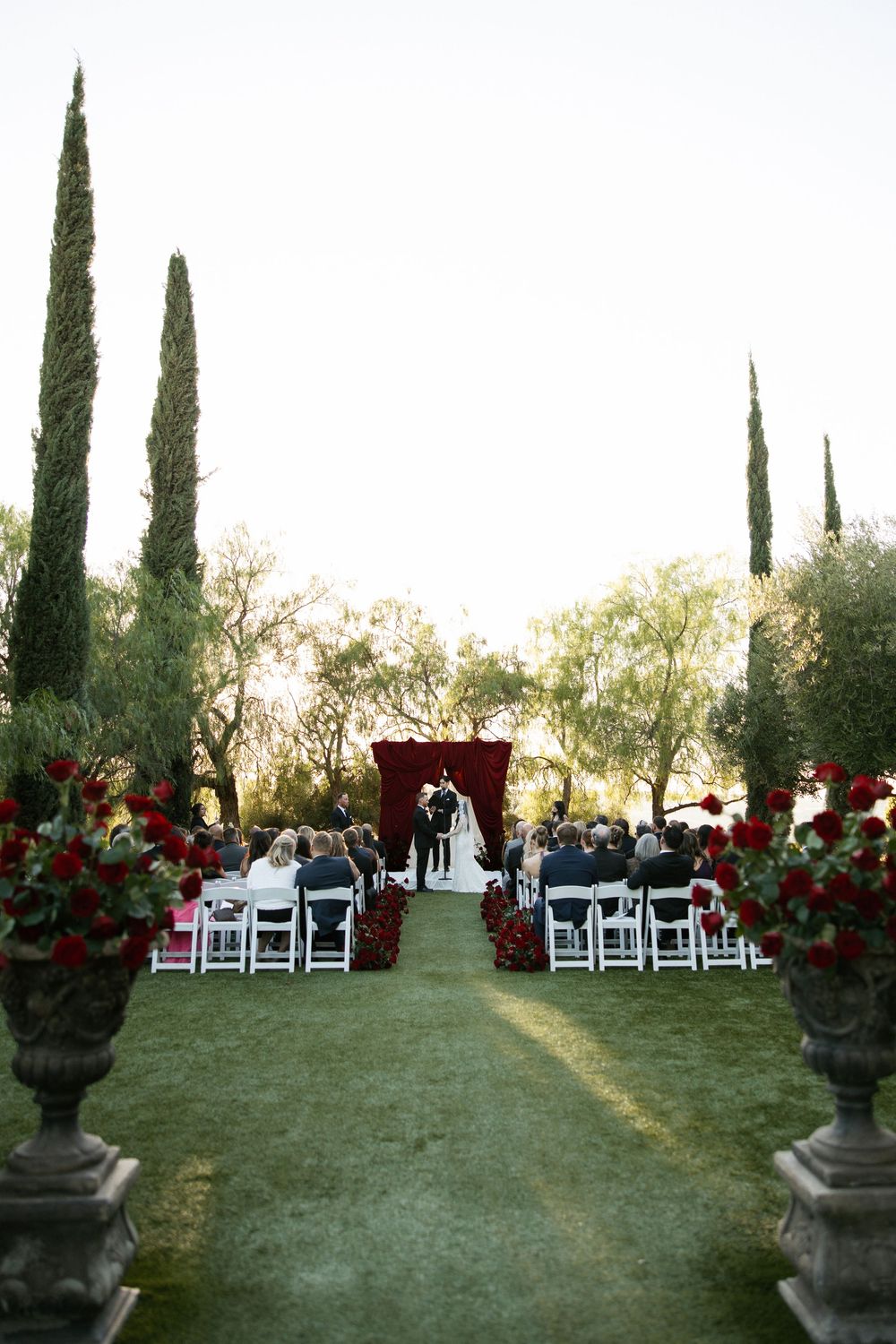Outdoor wedding ceremony with red floral arch, white chairs, and tall cypress trees on lush green lawn.