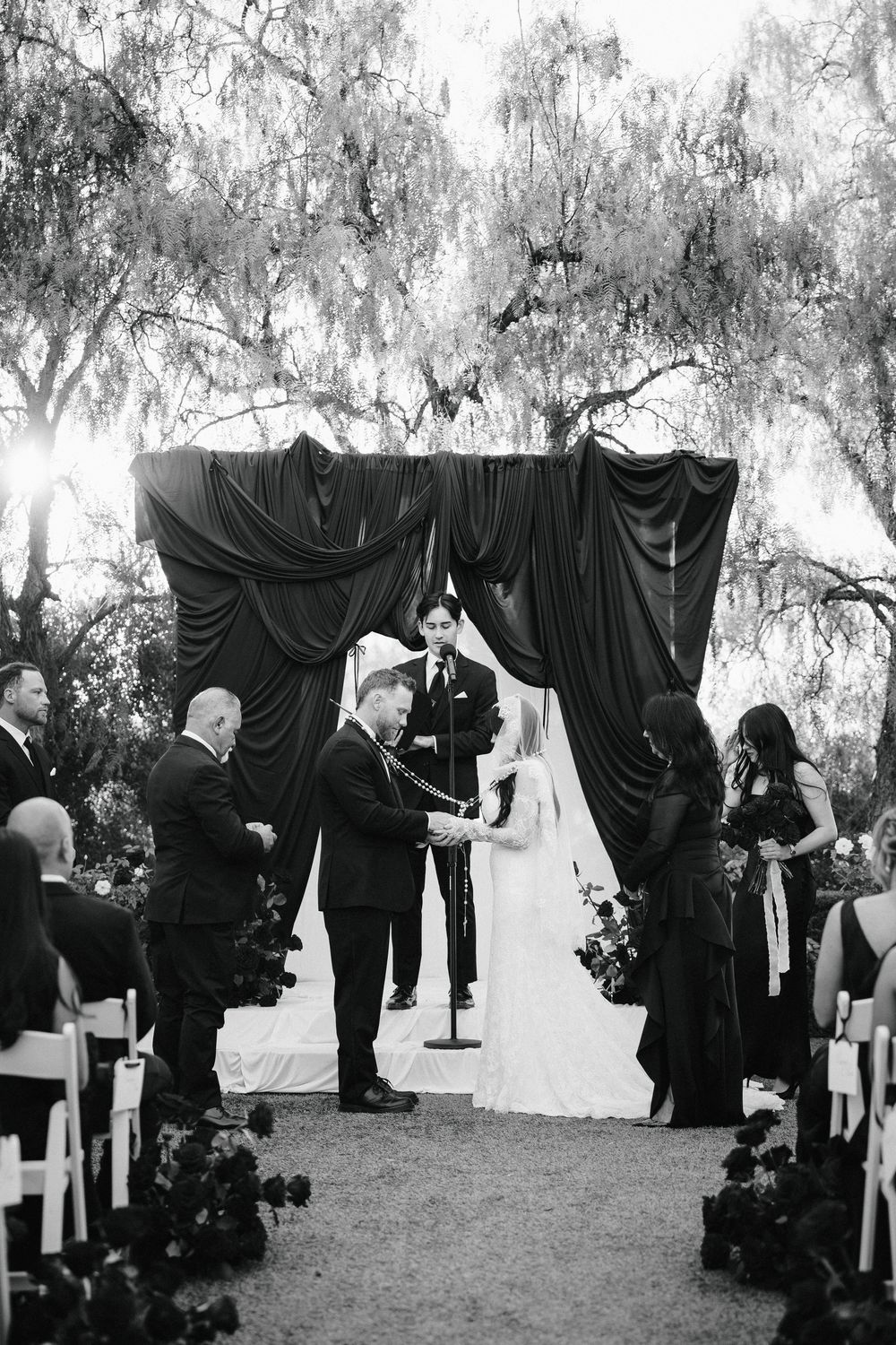 Outdoor wedding ceremony with red velvet draped arch, bride and groom exchanging vows surrounded by red roses.